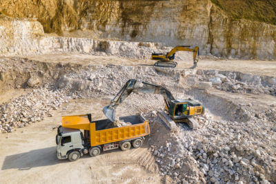 Ein Bagger belädt einen Muldenkipper mit Gestein in einem Steinbruch, ein weiterer Bagger steht im Hintergrund